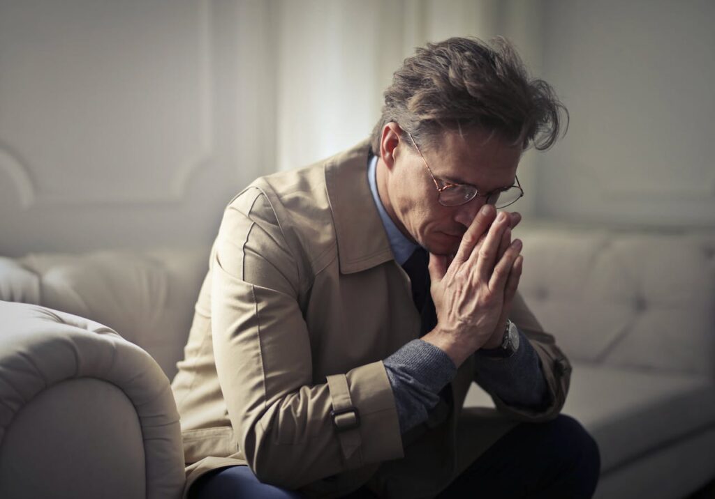 Side view of upset male entrepreneur in formal wear sitting on couch in modern living room and contemplating about financial problems while leaning on hands and looking down